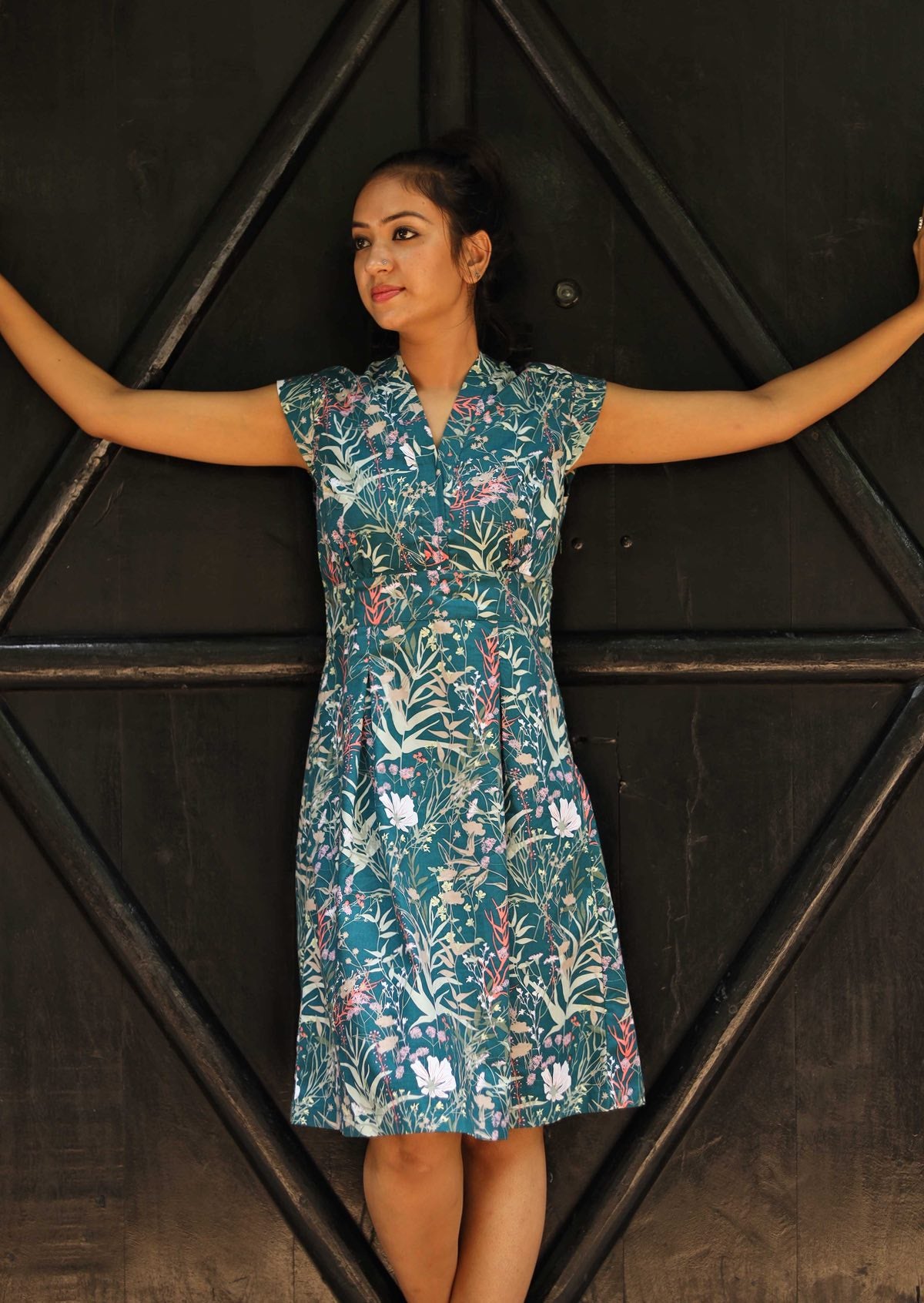 Woman wearing the Billie Dress Verdure, a structured cotton sundress with cap sleeves and a teal wildflower print in front of black door