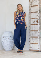 Woman wearing a sleeveless floral top and blue pants standing in a room with a large vase and wooden door.
