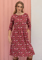 Model standing in front of pink background wears a knee length cotton dress with 3/4 sleeves, round neckline and pockets.
