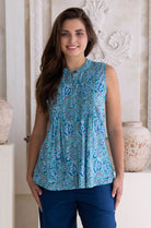 Woman wearing a aqua blue patterned sleeveless cotton blouse and dark blue pants standing in a decorative room.