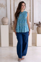 Woman wearing a blue patterned top and navy pants standing in a minimalistic room with white walls and light-colored floor.