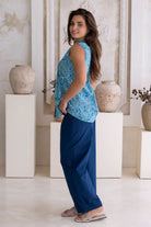 Woman wearing a aqua blue floral sleeveless cotton blouse and dark blue pants standing in a decorative room.