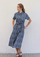 Woman poses in cotton blue based floral print shirt dress with fabric belt, with hand in pocket 
