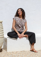 Woman sitting on a white concrete bench wears grey based floral print top featuring round neckline and cap sleeves 
