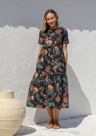 Woman wearing a 100% cotton, midlength black dress with botanical print featuring three tiered design, short sleeves, high round neckline and pockets