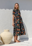 Woman wearing a relaxed fit printed black cotton dress with tiered design, standing next to a large vase against a white wall
