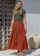 Woman standing in front of the wooden door wears 100% cotton button through  A line maxi skirt with belt loops and box pleats