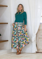 Woman wearing a green top and pastel spotty print button through skirt, leaning against a wall