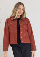Woman wearing a rust-colored jacket standing against a white wall.