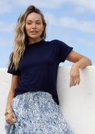 Woman wearing a navy t-shirt and blue floral skirt, leaning against a white wall