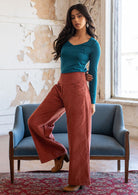 Woman wears burnt henna coloured corduroy wide legged pants with a side zip and pockets, paired with a teal long sleeve rayon top