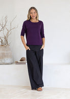 Woman wearing a purple relaxed fit top with 3/4 sleeves and black pants standing in a minimalistic room.