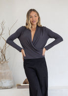 Woman wearing a dark grey long sleeve crossover top with pleats at the front of the shoulders with black pants