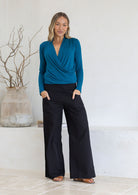 Woman wearing a long sleeve teal crossover top and black pants standing against a white wall.