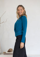 Woman wearing a teal long sleeve top with gathers where the top crosses over at the front and black pants