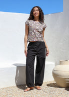 Woman standing in sun wears a floral cotton top with wide leg black cotton pants with elastic either side of waist and pleated ankles