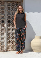 Woman wearing a black sleeveless top and floral wide leg cotton pants with hands in pockets, standing in front of a textured wooden door.