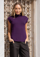Woman wearing a purple sleeveless top with a high neckline and black pants standing against a wooden wall.