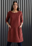 Woman wearing rust coloured corduroy tunic with sleeves over navy blue leggings and boots with hands in pockets 