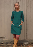 Woman with blond hair wearing Teal corduroy dress over pink leggings with 3/4 sleeves with hands in pockets 