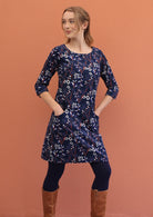Woman wears  round neck, 3/4 sleeve corduroy dress with hands in pockets, paired with navy blue leggings and boots