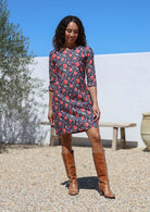 Woman wearing a navy cotton floral dress and brown boots standing outdoors with a white wall and tree in the background.