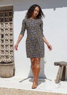 Woman wearing a black floral cotton patterned dress and brown boots standing against a white wall.