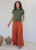 Model posing in wide leg rust cotton pants with drawstring waist tie and elastic at back of the waist.