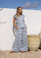Woman in a white based blue floral cotton pant wide legged against a white wall.