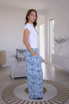 Woman standing sideways wearing a white basic top and white based blue floral pants standing in a modern living room.