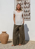 Woman wearing a white cotton top and olive green wide leg cotton pants standing in front of a white wall with a wooden window
