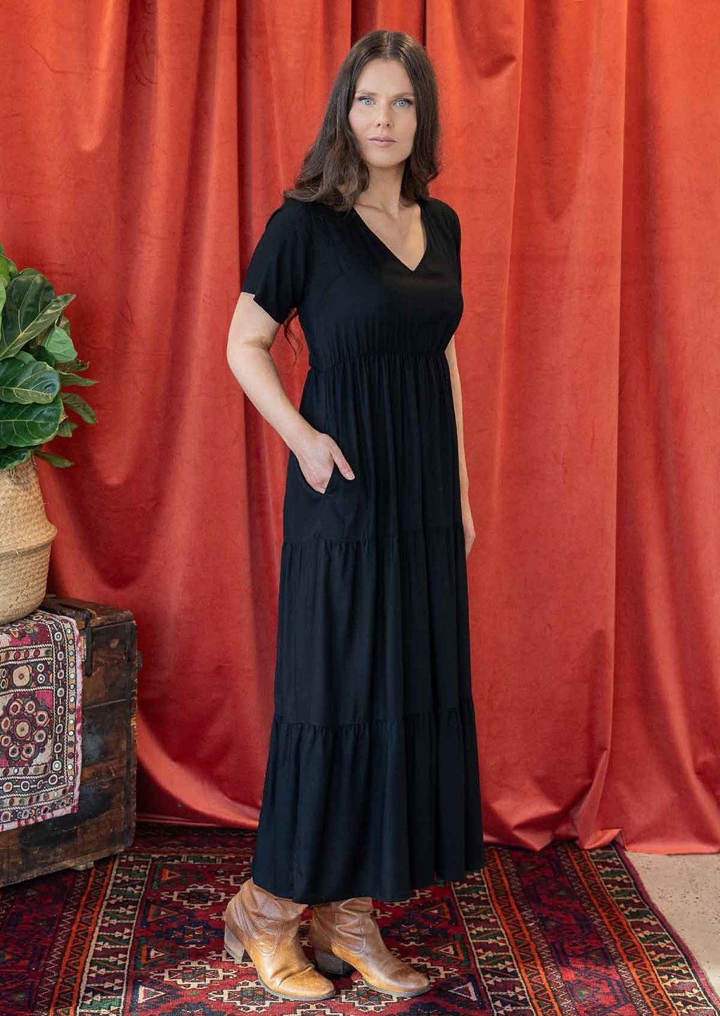 Woman in a black rayon maxi dress with a V-neckline and short sleeves, standing against a salmon coloured curtain