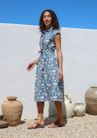 Model stands sideways wearing a floral printed blue shirt style cotton dress with collar, buttoned bodice, pockets and cap sleeves