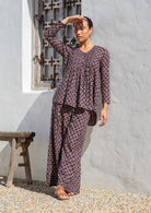 Model standing in sun wears long sleeve button through top with V-neck and small pleats across chest that leads into A line flare at the base.