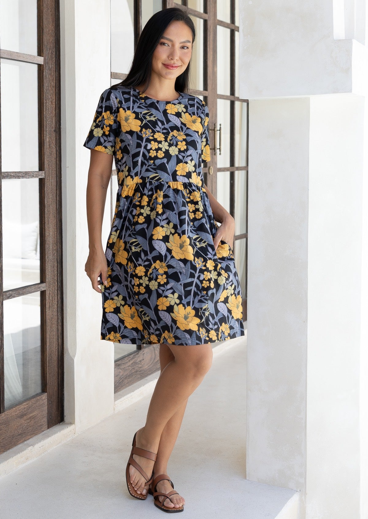 Woman stands with hand in pocket wearing a 100% cotton relaxed fit dress with round neckline and short sleeves in black based retro floral print