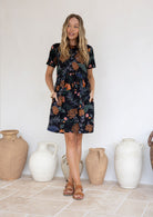 Model wears short sleeved, above knee 100 percent cotton dress with high round neckline and both the hands in pocket.