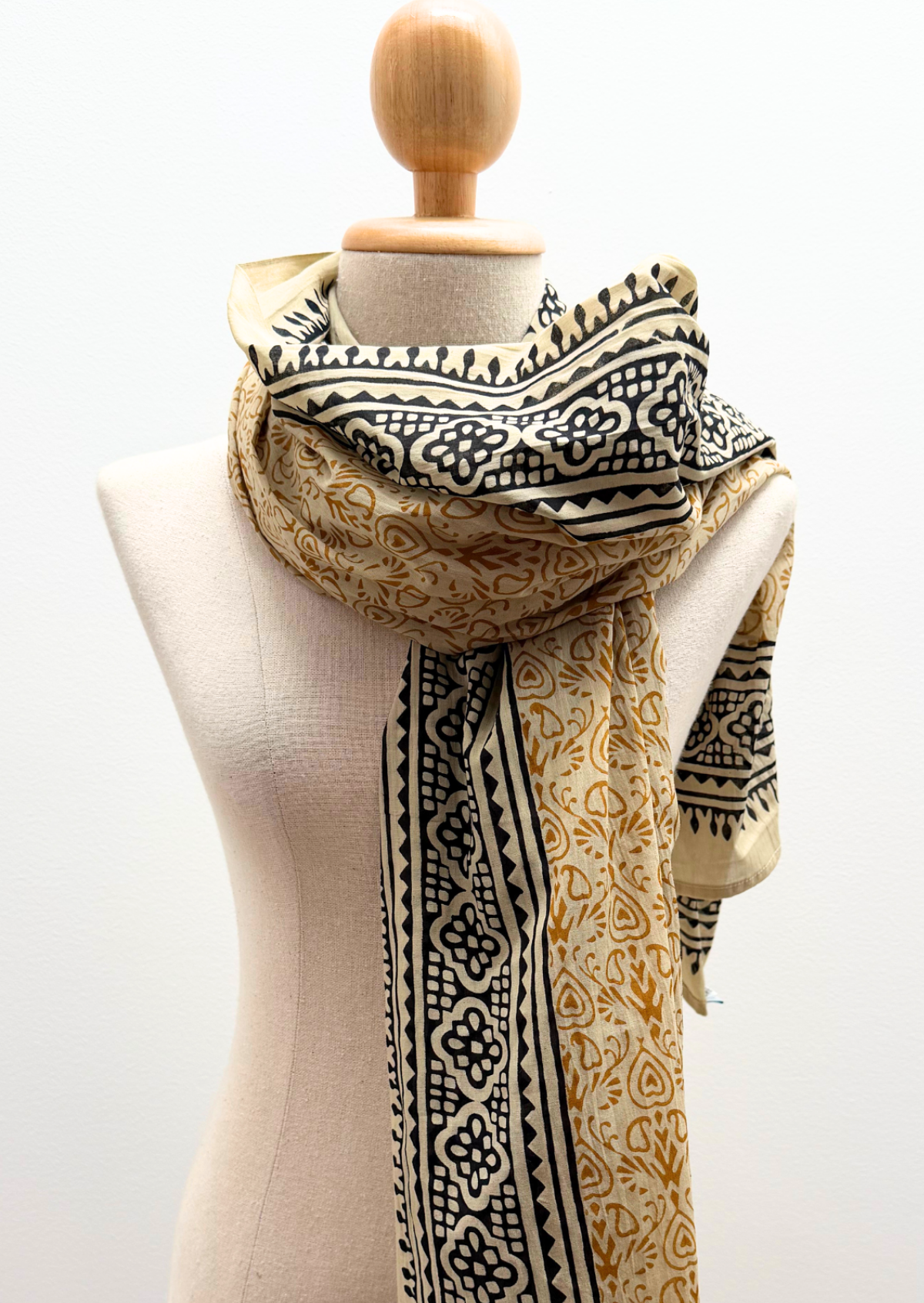 A patterned scarf draped on a cream-colored dress form mannequin against a plain white background. The scarf features a beige base with black geometric borders and a golden-yellow paisley pattern.