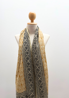 A lightweight scarf with a black and gold block-printed geometric pattern, draped on a cream-colored mannequin torso against a plain white background.