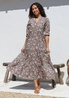 Woman standing against the white wall wears 3/4 sleeved cotton tiered maxi dress with mandarin collar and buttoned bodice 