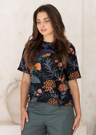 Woman wearing a loose fit black floral with green pants against a white wall.