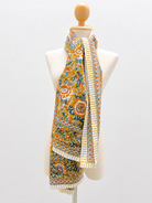 A patterned scarf draped on a cream-colored mannequin torso against a light gray studio background. The scarf features a dense floral and paisley print in mustard yellow, orange, green, and blue, with cream and orange striped borders and fringe.