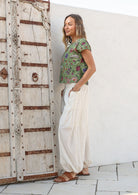 Woman wears lightweight white cotton harem pants paired with a green floral top and sandals, with hands in pockets