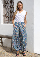 Woman wearing a white top and blue floral pants with wide elastic waist and ankles standing in front of a white wall with a wooden bench.