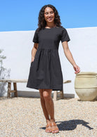 Woman wearing a black loose fit above knee length cotton dress with short sleeves and high round neckline