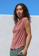 Woman wearing a pink sleeveless top with draped neck against a white wall with blue sky.