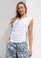 Woman wearing a white sleeveless top with pleats at each shoulder paired with floral pants against a light background