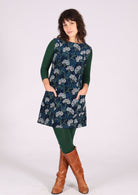 Woman poses in floral print cotton corduroy sleeveless tunic layered with dark green leggings, long sleeve top and boots