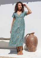 Woman wears 100% cotton midlength dress in sage green, buttoning through to the knee with V neckline, pockets and shoestring tie 