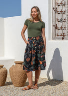 Woman wearing a knee length, box pleated cotton skirt with button front, belt loops and pockets in black based botanical print