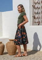 Model poses with hands in pockets wearing a knee length black cotton skirt featuring a lovely botanical print with belt loops, box pleats and elastic back waist